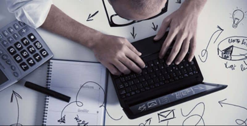 Overhead view of a person's hands typing on a laptop keyboard surrounded by a calculator, notebook, pen, and hand-drawn notes